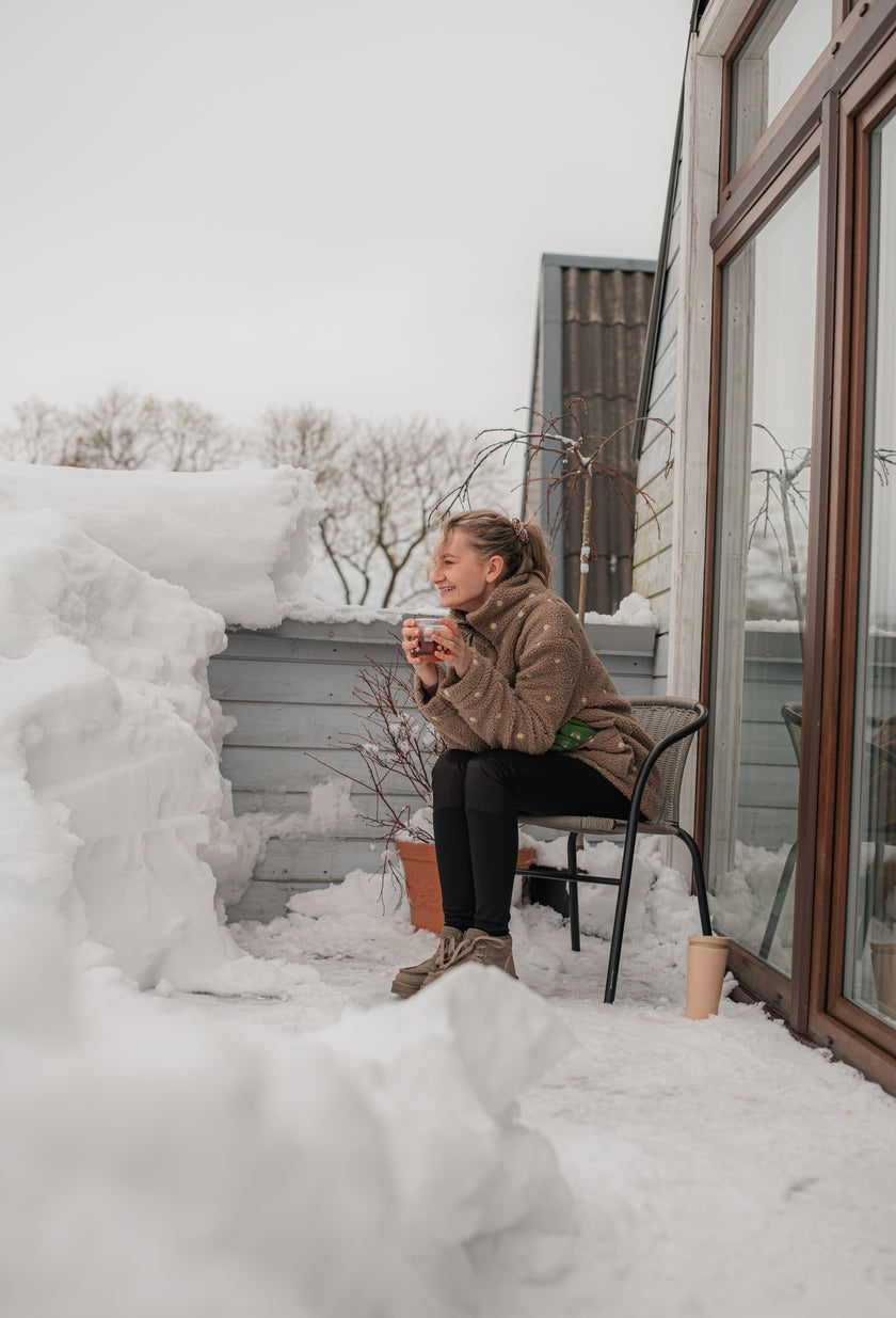 Moteris su gėlėtu teddy paltuku sėdi žiemos terasoje tarp sniego — laiko puodelį ir šypsosi